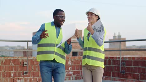 Young-and-attractive-African-American-cheerful-man-and-happy-Caucasian-woman,-builders-in-hardhats-dancing-on-the-roof-of-the-building-at-the-construction-site.-Outside.